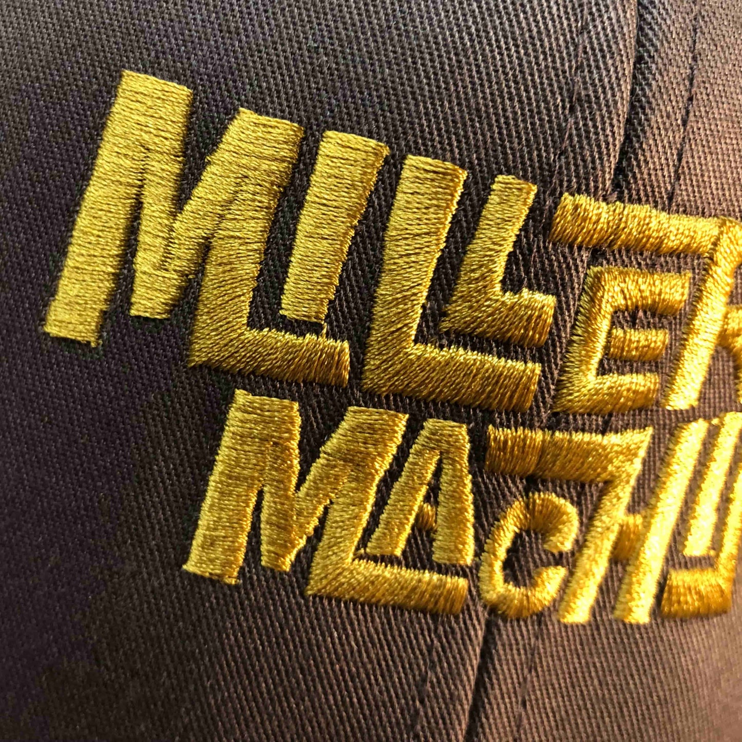 Closeup photo of gold embroidery on the Miller Machine Trucker Cap in brown.