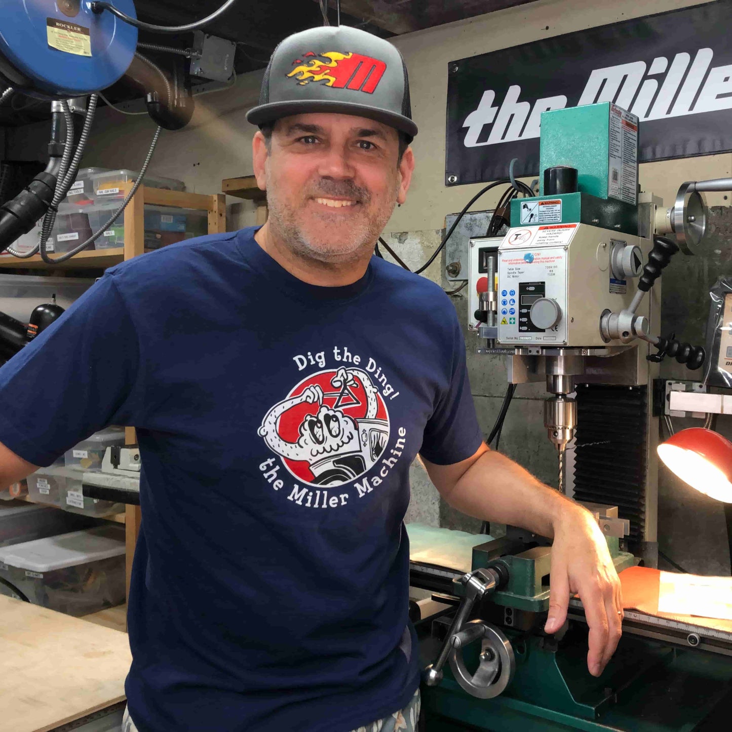 Photo of Billy Miller wearing a Miller Machine 5 panel trucker cap with a Flaming M in red and yellow. Cap is a grey color.