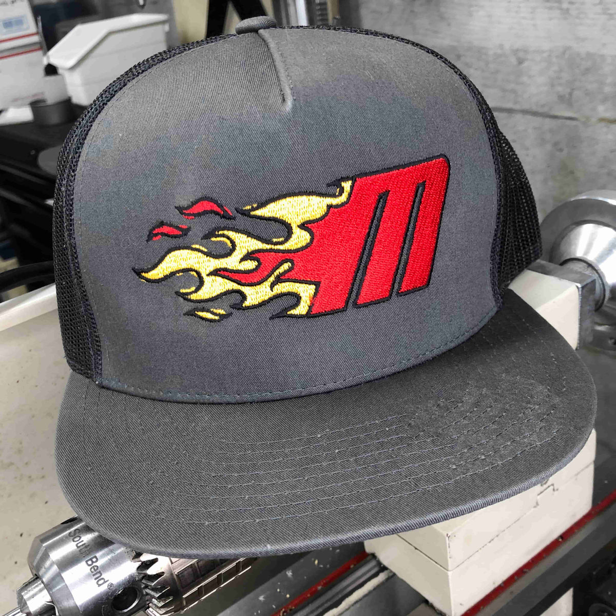 Photo of Miller Machine 5 panel trucker cap with a Flaming M in red and yellow. Cap is a grey color.