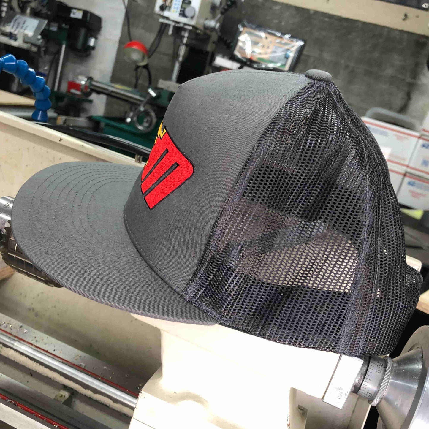 Side view of Miller Machine 5 panel trucker cap with a Flaming M in red and yellow. Cap is a grey color.